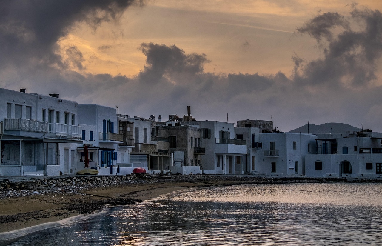 White buildings lined along a seaside at sunset,with calm water reflecting the light & dramatic clouds in the sky.